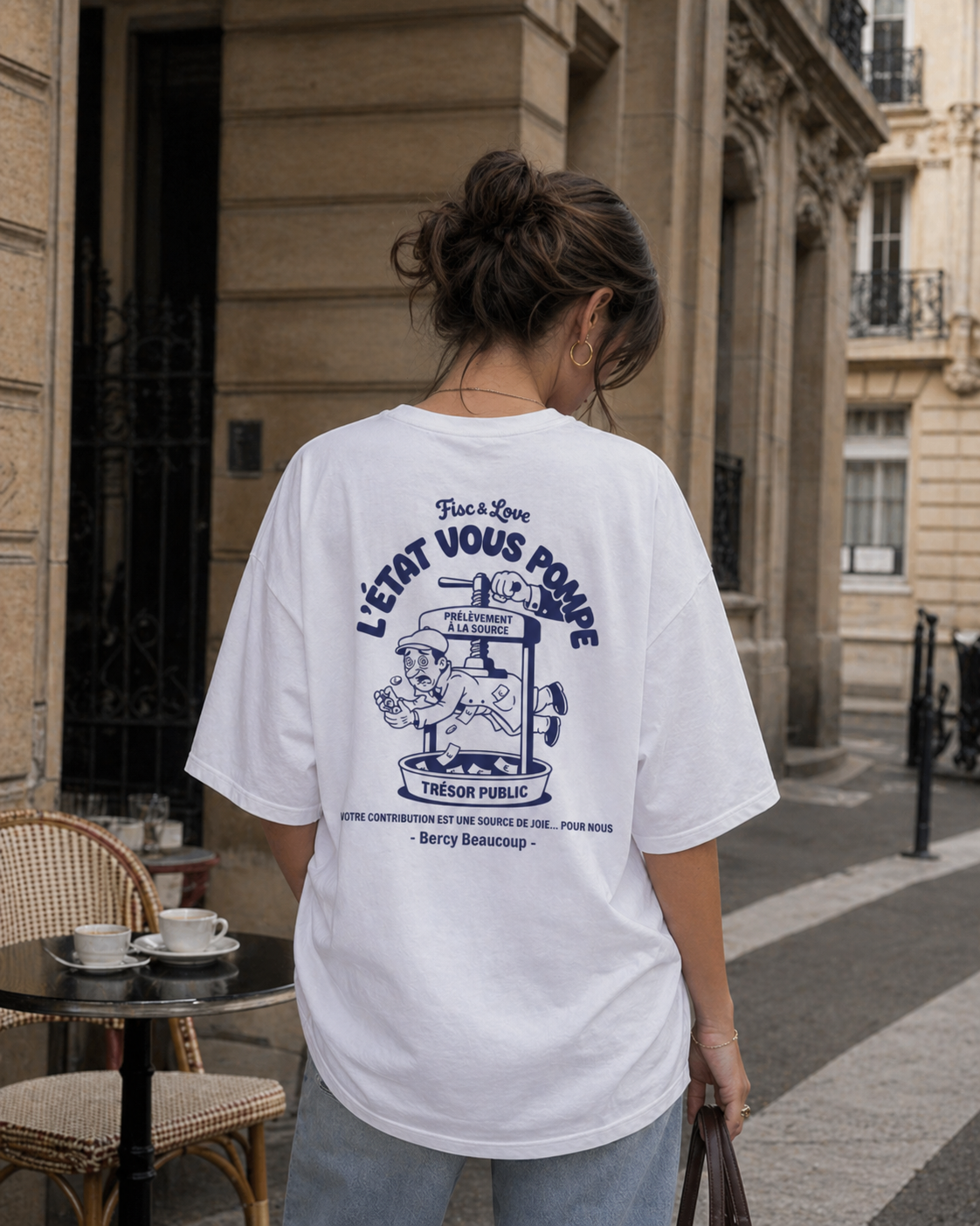 Vue de dos d'une femme avec un chignon, debout à une terrasse de café à Paris. Elle porte un t-shirt oversize blanc avec une illustration satirique bleu marine. Le dessin représente un homme écrasé dans un pressoir intitulé « PRÉLÈVEMENT À LA SOURCE », avec l'argent tombant dans une cuve « TRÉSOR PUBLIC ». On lit en haut « L'ÉTAT VOUS POMPE » et en bas le message ironique : « VOTRE CONTRIBUTION EST UNE SOURCE DE JOIE... POUR NOUS. - Bercy Beaucoup - ». L'arrière-plan montre des immeubles haussmanniens et un