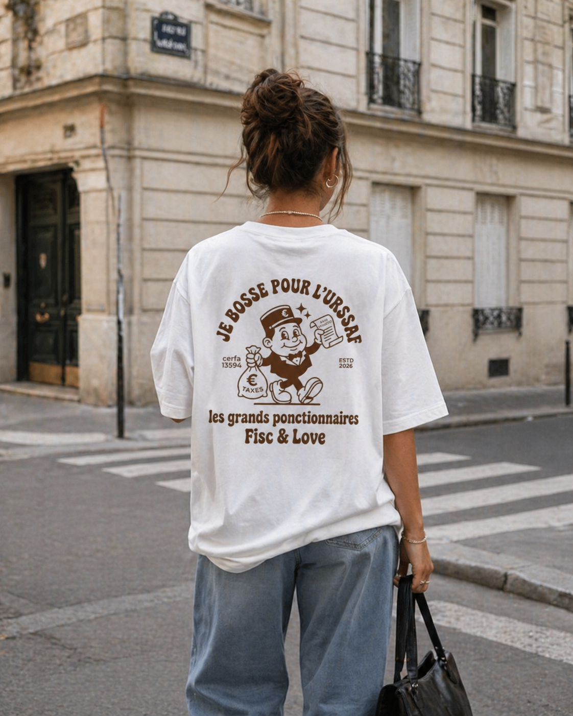 Photographie de dos d'une femme portant un t-shirt blanc oversize dans une rue de Paris. Le dos du t-shirt arbore une illustration marron style dessin animé d'un homme en costume tenant un sac d'argent et un document, entouré des textes "JE BOSSE POUR L'URSSAF", "cerfa 13594", "EST. 2023", "les grands fonctionnaires" et "Fisc & Love". La femme porte un jean bleu et tient un sac noir. Des immeubles haussmanniens et un passage piéton sont visibles en arrière-plan