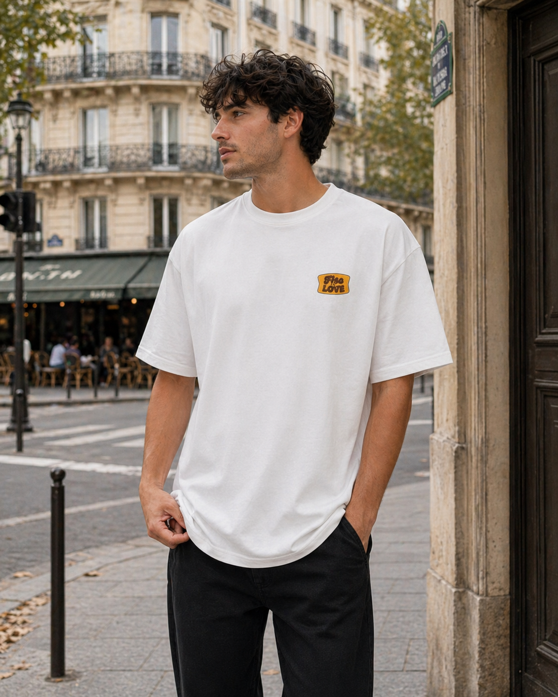 Portrait d'un homme de profil portant un t-shirt blanc oversize avec un petit logo jaune sur la poitrine et un pantalon noir. Il se tient debout dans une rue de style parisien devant un immeuble haussmannien et une terrasse de café en arrière-plan