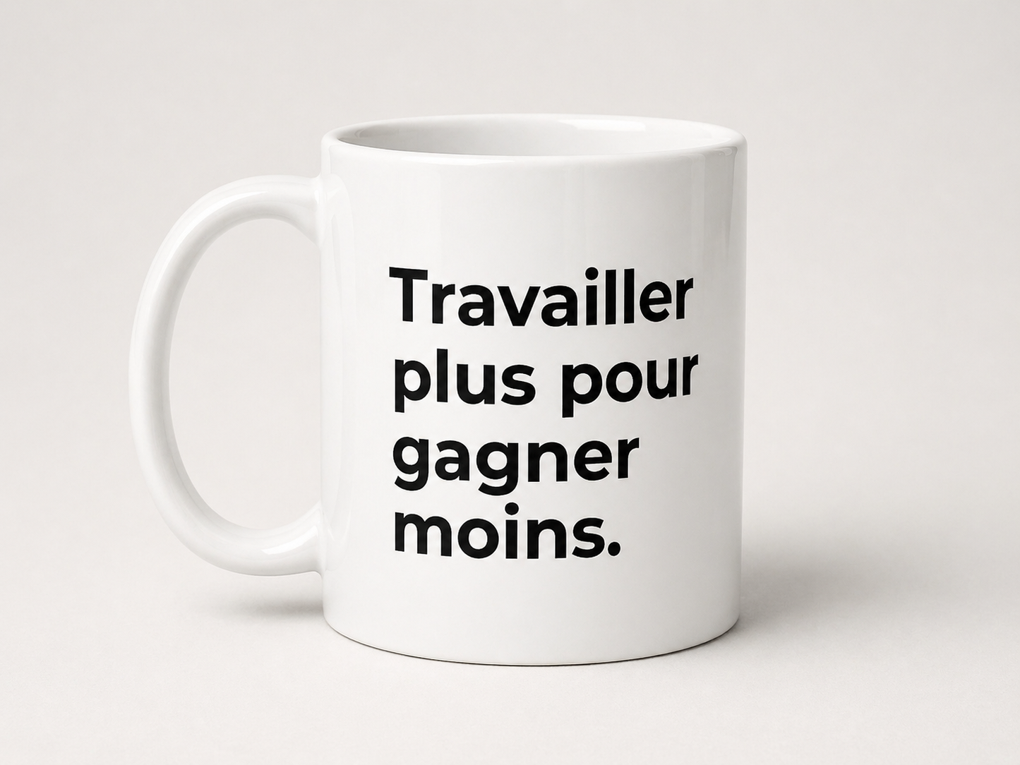 Photographie de studio d'un mug en céramique blanche sur un fond neutre clair. Le mug affiche une inscription en lettres noires grasses et sans empattement sur quatre lignes : « Travailler plus pour gagner moins. ». Le texte détourne de façon satirique un célèbre slogan politique français. L'éclairage est doux, mettant en valeur la forme cylindrique épurée de la tasse