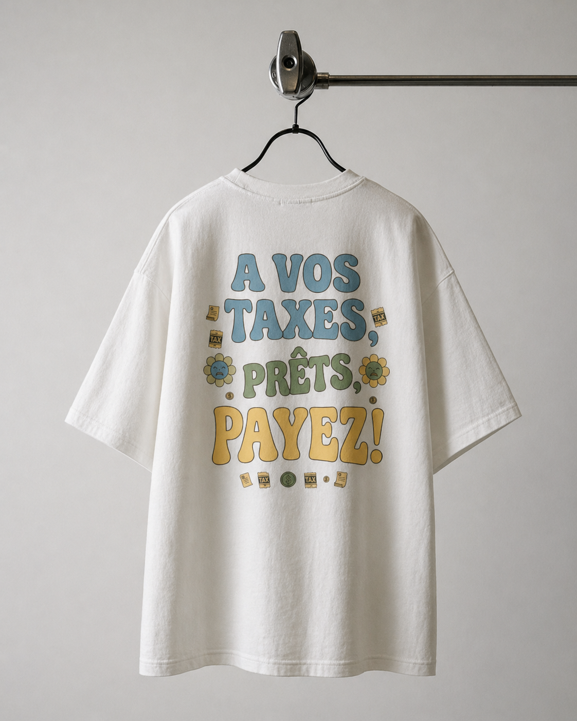 Dos d'un t-shirt blanc oversize avec l'inscription graphique 'À VOS TAXES, PRÊTS, PAYEZ !' en lettres colorées bleues, vertes et jaunes, entourées de petites fleurs et de motifs de formulaires de taxes."