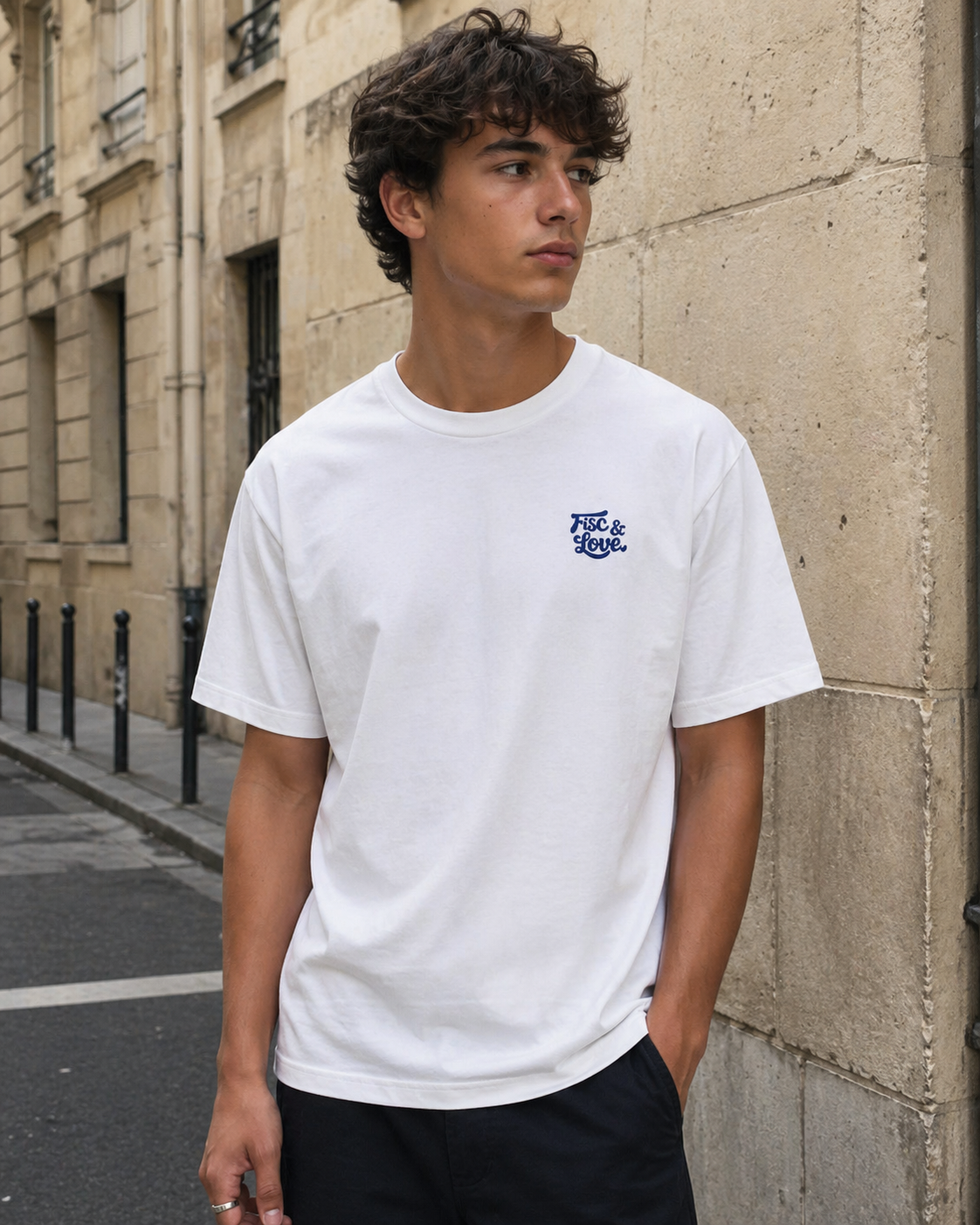 Photographie en plan moyen d'un jeune homme aux cheveux bouclés, posant dans une rue parisienne pavée devant un mur de pierre. Il porte un t-shirt blanc oversize avec un grand graphisme détaillé en broderie bleu foncé sur la poitrine. Le graphisme est une illustration de style dessin animé vintage centrée sur le personnage de « L'Aspirateur Fiscal », qui utilise un aspirateur pour créer un vortex aspirant des billets de banque, des citoyens, des buissons « TAX » et « URSSAF », et même la Tour Eiffel. Le tex