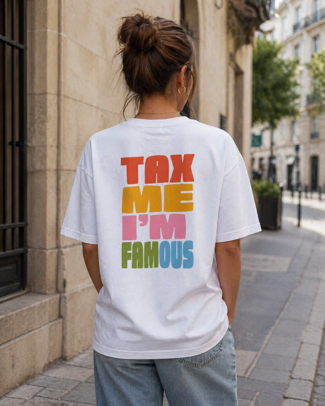 Photo de style de vie prise de dos montrant une femme avec un chignon portant un t-shirt blanc oversize. Le dos du t-shirt affiche le message satirique "TAX ME I'M FAMOUS" écrit en grandes lettres colorées (rouge, jaune, rose et bleu). Elle se tient dans une rue pavée ensoleillée avec des bâtiments classiques en arrière-plan.