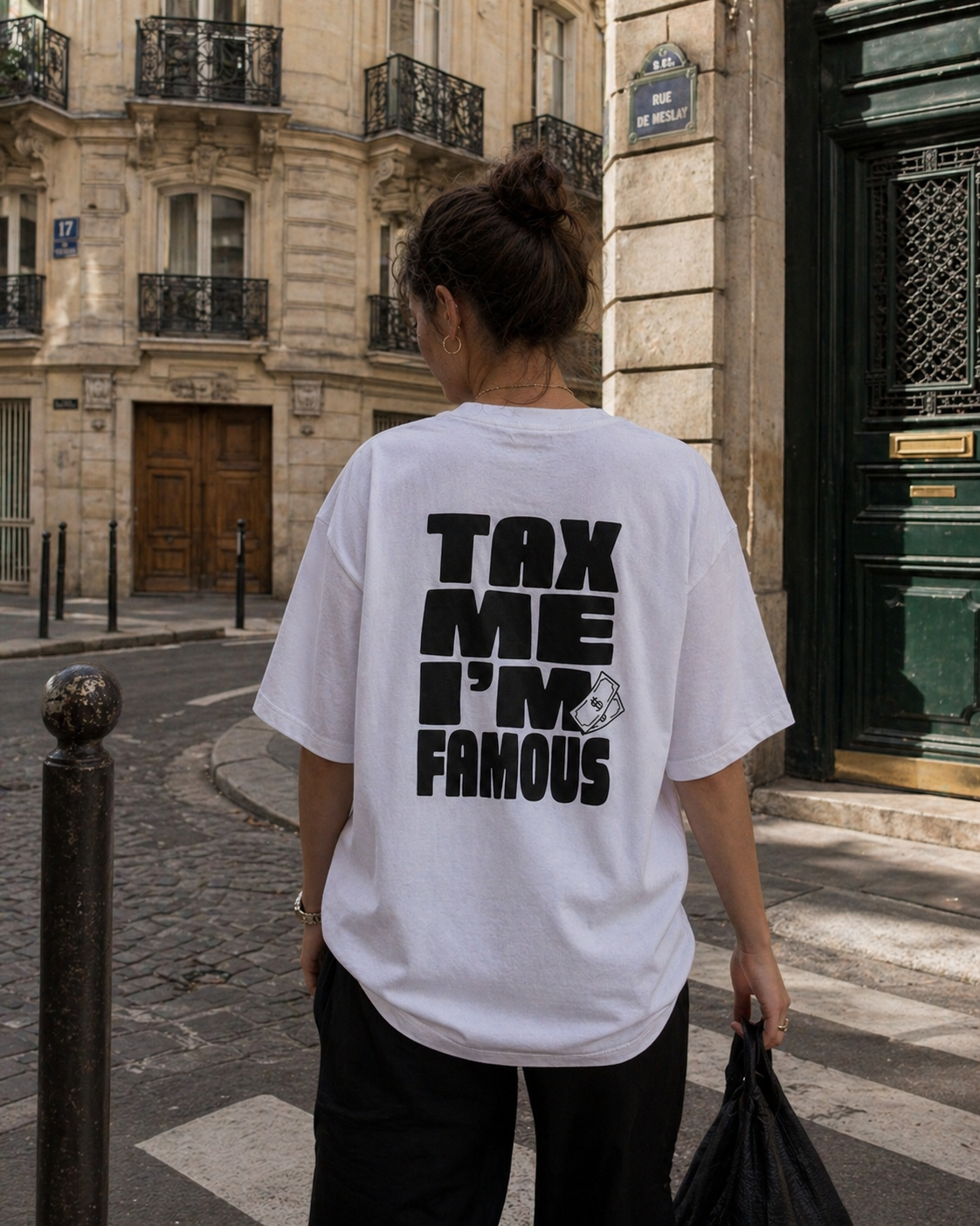 Photo de style de vie de dos d'une femme portant un t-shirt blanc oversize avec un petit logo multi-coloré "FISC & LOVE" sur la poitrine. Elle se tient dans une rue pavée de Paris, avec un panneau "Rue Saint-André-des-Arts" visible. L'arrière-plan montre des immeubles haussmanniens classiques sous une lumière naturelle