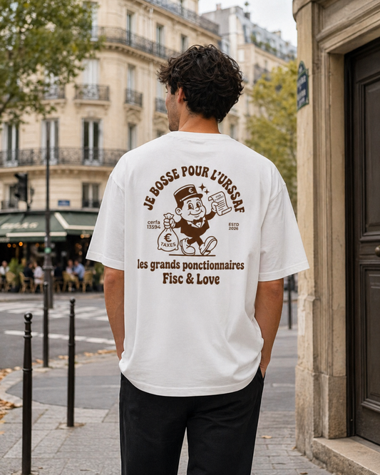 Photo de style de vie de dos d'un homme portant un t-shirt blanc oversize dans une rue de Paris. Le dos affiche une illustration satirique marron de style vintage avec le texte "JE BOSSE POUR L'URSSAF" et un personnage de cartoon tenant un sac de taxes. On aperçoit des immeubles haussmanniens et une terrasse de café en arrière-plan