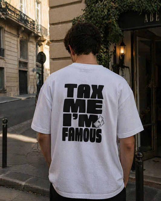 Photo de dos d'un homme portant un t-shirt blanc oversize dans une rue urbaine. Le dos est imprimé avec le message "TAX ME I'M FAMOUS" en lettres capitales noires imposantes, accompagné d'une petite illustration de billets de banque. L'arrière-plan montre une façade en pierre classique et l'entrée d'un établissement.