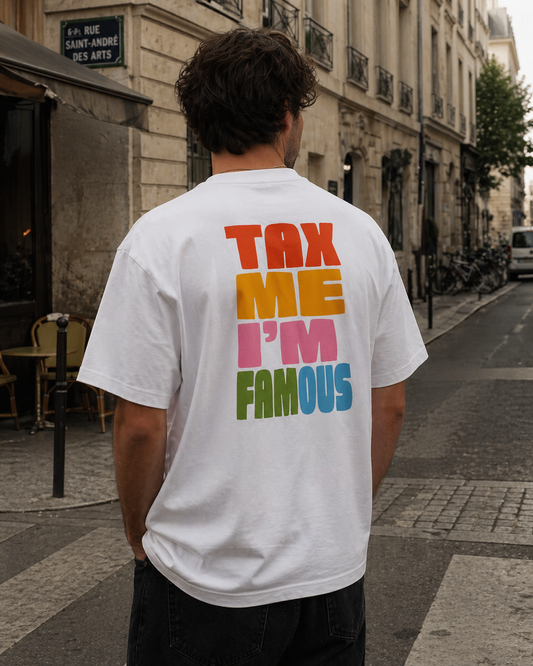 Photo de dos d'un homme portant un t-shirt blanc oversize avec un grand imprimé coloré "TAX ME I'M FAMOUS". Il se tient dans une rue pavée typique de Paris, près d'un panneau "Rue Saint-André-des-Arts" et d'arbres. Ses mains sont dans ses poches de pantalon noir