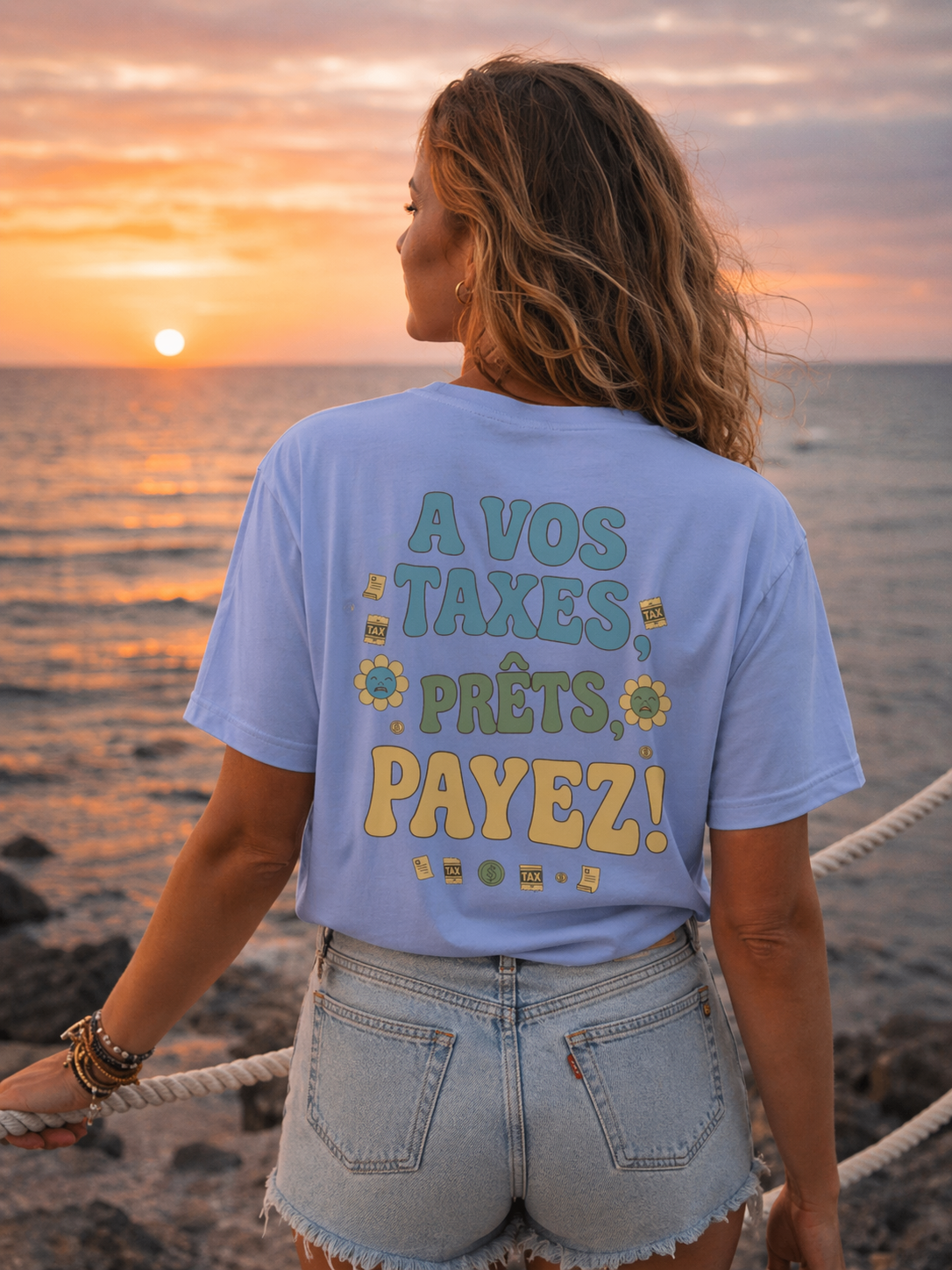Gros plan sur le design graphique du t-shirt Fisc & Love affichant le slogan satirique 'À VOS TAXES, PRÊTS, PAYEZ !'. Le visuel présente des lettres colorées de style rétro, des illustrations de billets de banque et des fleurs printanières, emblématique de l'univers décalé de Fisc & Love